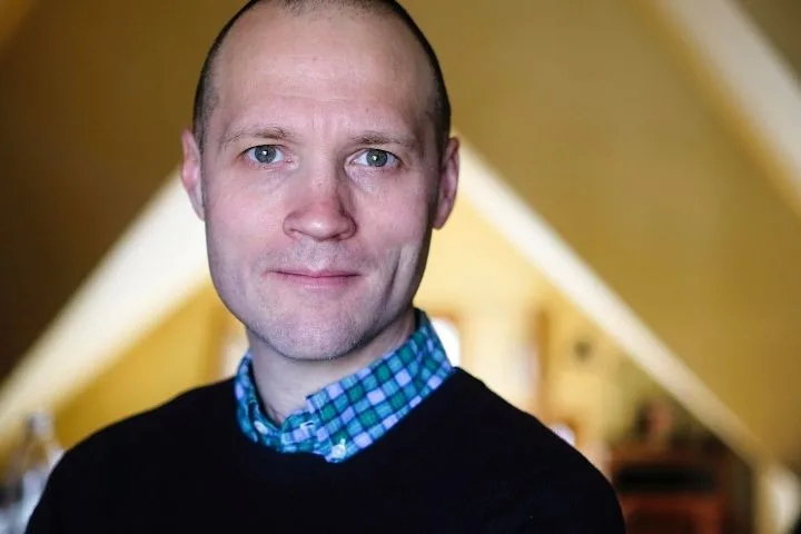 A man with blue eyes and a shaved head, wearing a dark sweater over a blue and green plaid shirt, stands indoors with blurred, warm-toned background and slanted ceiling beams behind him.