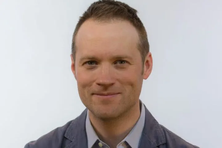 A man with short brown hair, fair skin, and blue eyes smiles slightly at the camera. He is wearing a gray collared shirt and a gray blazer. The background is plain and white.