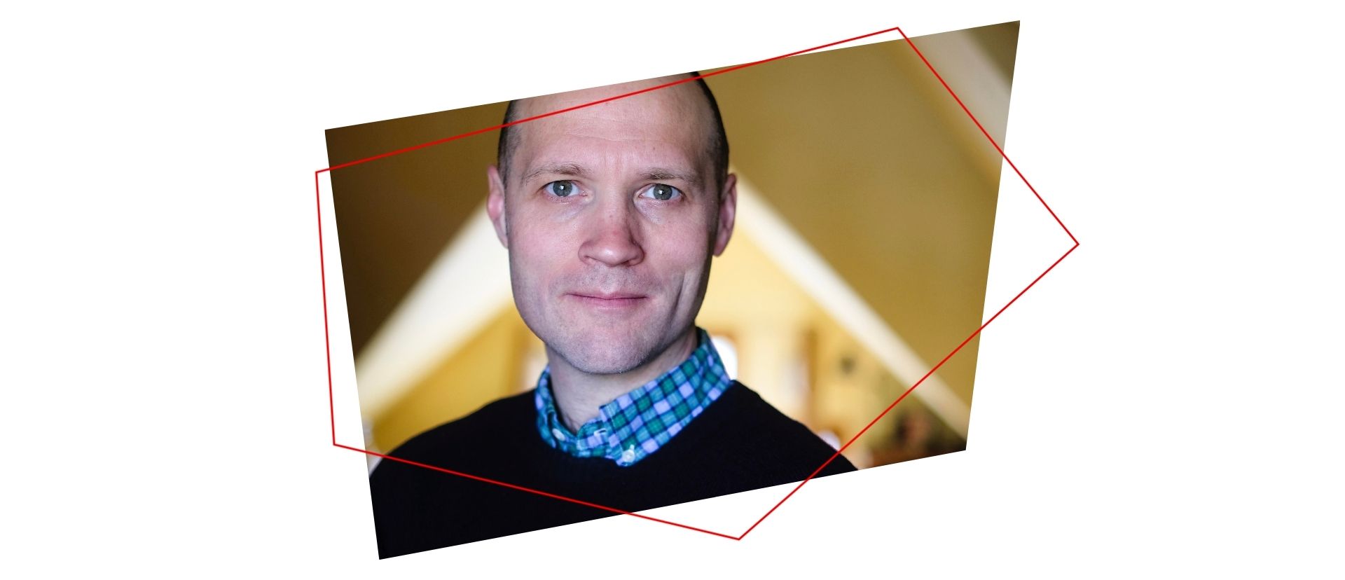 A man with short hair and blue eyes, wearing a blue plaid shirt and a dark sweater, stands indoors with a blurred background. A red geometric outline frames the photo at an angle.
