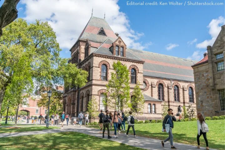 Students walk and gather on a sunny day in front of a historic stone building with arched windows and a steep roof on a college campus, surrounded by green trees and grass.