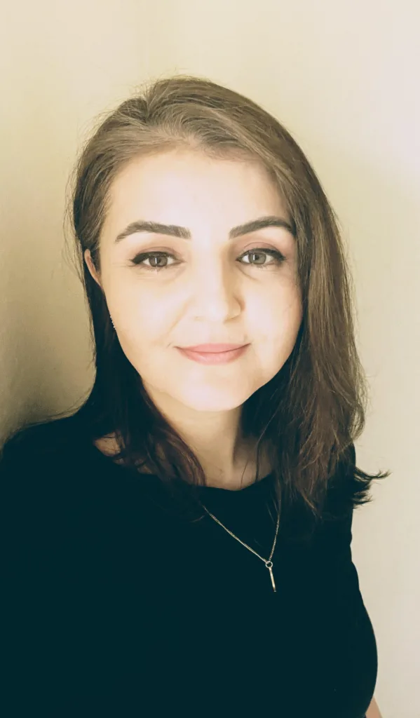 A woman with straight, shoulder-length brown hair, wearing a black top and a delicate necklace, stands against a light-colored background, smiling softly at the camera.