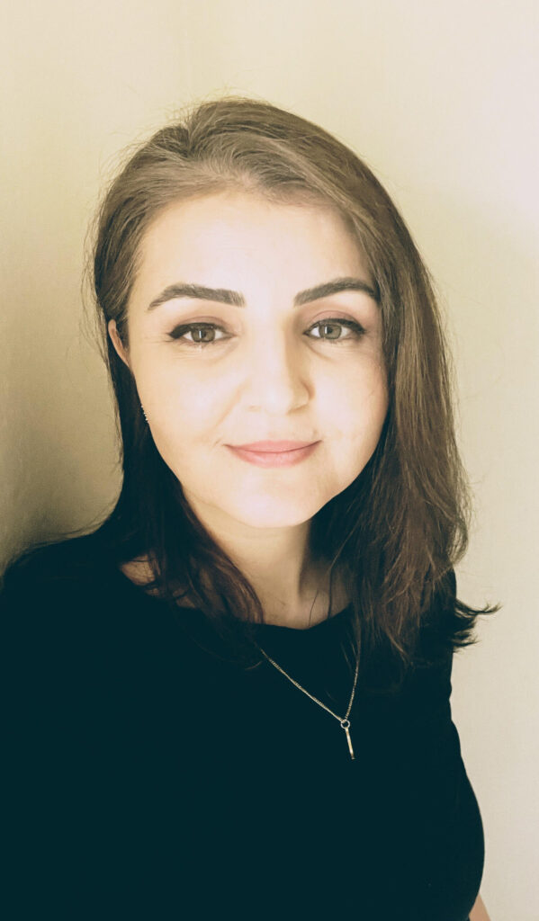 A woman with straight, shoulder-length brown hair, wearing a black top and a delicate necklace, stands against a light-colored background, smiling softly at the camera.