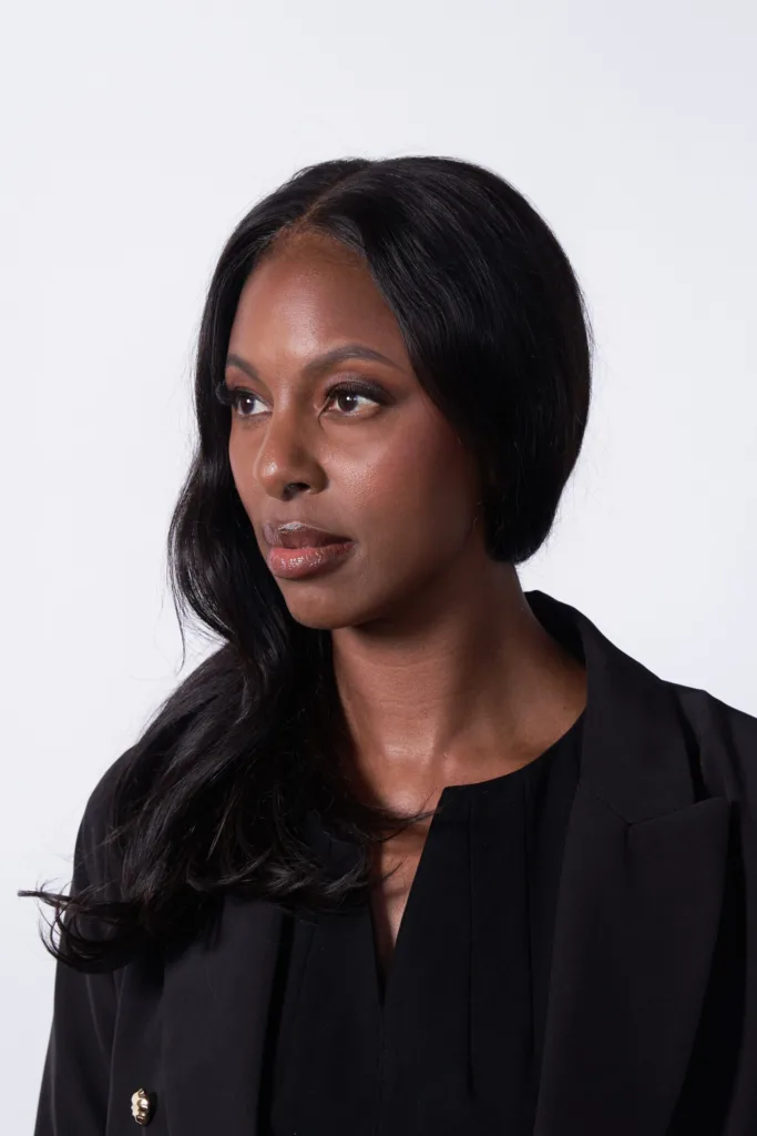 A woman with long, wavy black hair wearing a black blazer and black top looks thoughtfully to her right against a plain white background.