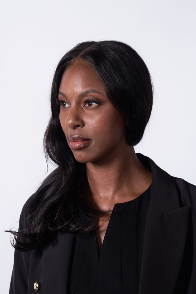 A woman with long, wavy black hair wearing a black blazer and black top looks thoughtfully to her right against a plain white background.