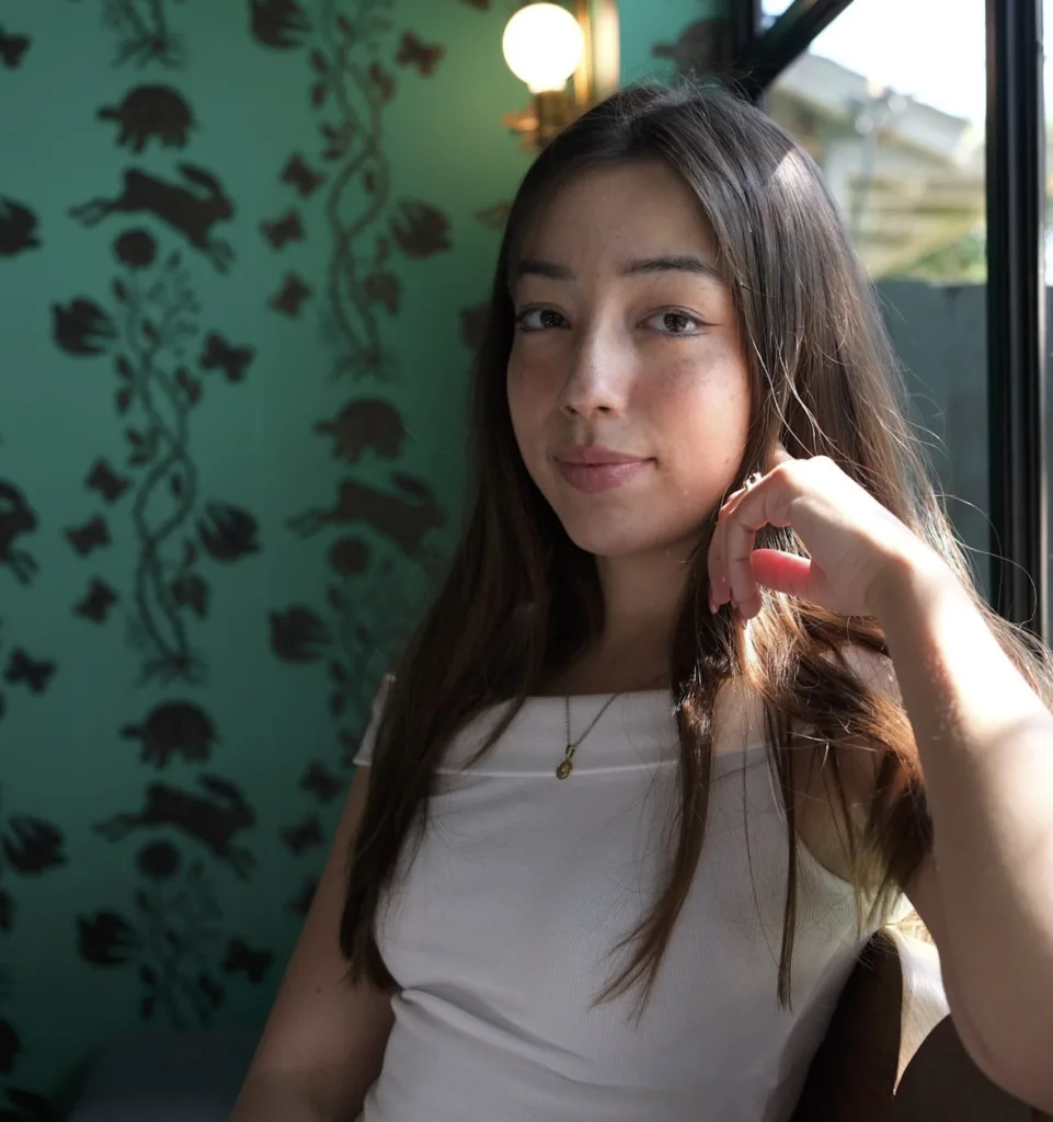 A young woman with long brown hair sits by a window, resting her hand near her face. She wears a white top, a gold necklace, and gazes calmly at the camera. The background features teal wallpaper with a dark floral pattern.