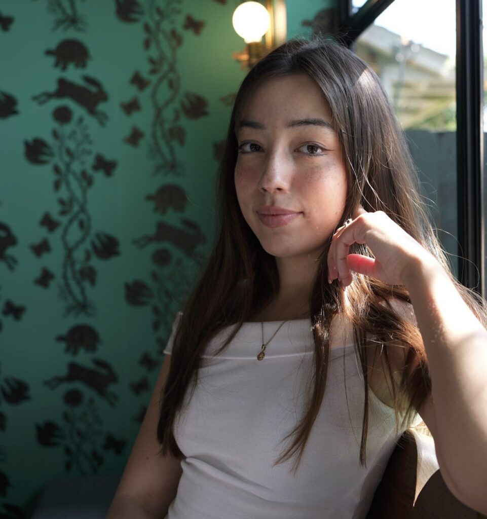 A young woman with long brown hair sits by a window, resting her hand near her face. She wears a white top, a gold necklace, and gazes calmly at the camera. The background features teal wallpaper with a dark floral pattern.