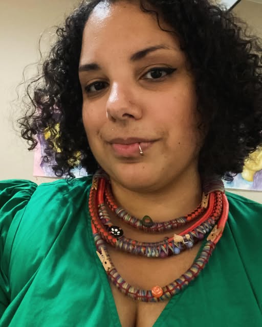 A person with curly hair, wearing a green top and layered colorful bead necklaces, looks at the camera with a slight smile. They have a pierced lip and winged eyeliner, and the background is softly blurred.
