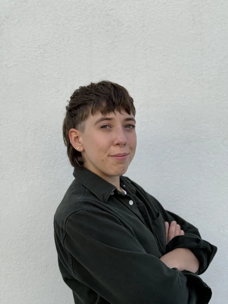 A person with short brown hair and a mullet hairstyle stands in front of a white wall, arms crossed, wearing a dark button-up shirt and looking at the camera with a slight smile.