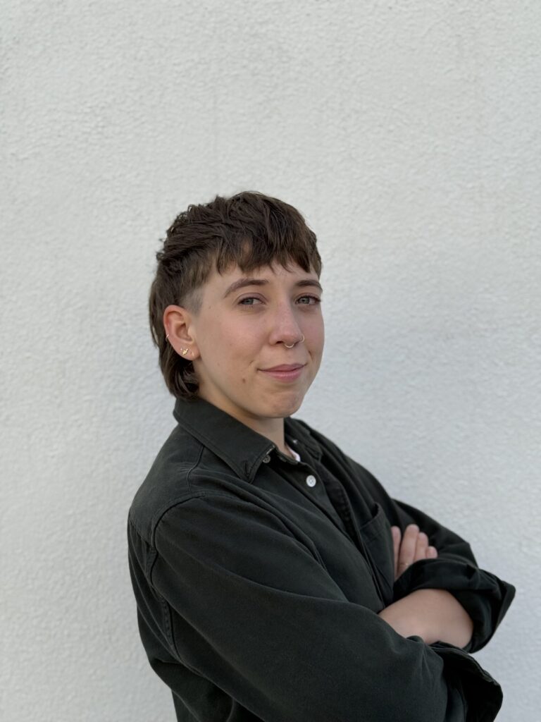 A person with short brown hair and a mullet hairstyle stands in front of a white wall, arms crossed, wearing a dark button-up shirt and looking at the camera with a slight smile.