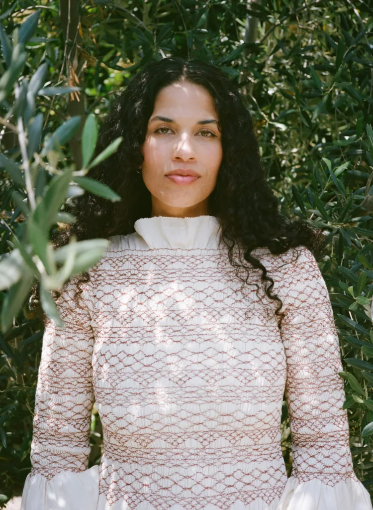 A woman with long, curly black hair stands outdoors among green leafy branches, wearing a high-necked, textured light-colored dress with red patterns. She looks directly at the camera with a calm expression.