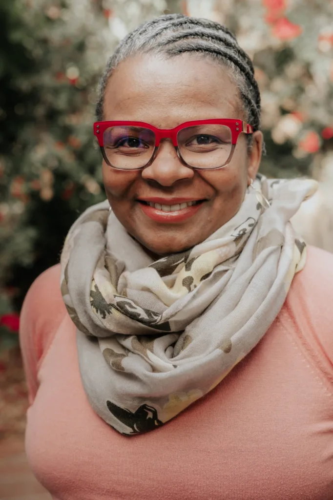A woman with braided hair, wearing red glasses, a patterned scarf, and a pink top, smiles at the camera. The background is blurred with greenery and red flowers.