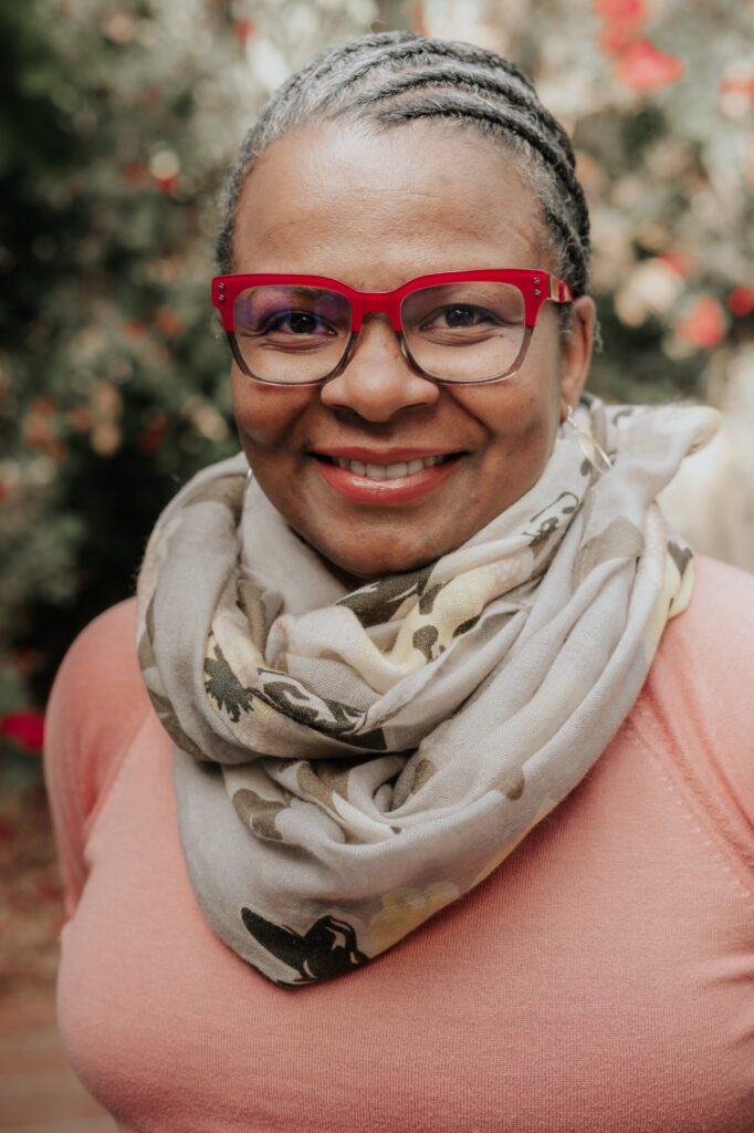 A woman with braided hair, wearing red glasses, a patterned scarf, and a pink top, smiles at the camera. The background is blurred with greenery and red flowers.