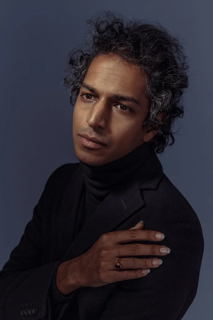 A person with wavy, graying hair wearing a black turtleneck and blazer poses against a dark blue background, looking thoughtfully into the distance with one hand resting on their shoulder.