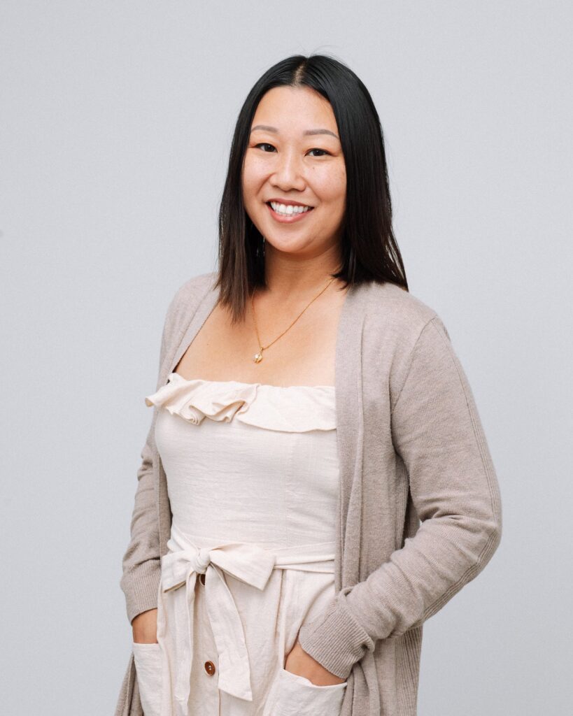 A woman with straight dark hair smiles, wearing a light beige dress with a matching cardigan and hands in her pockets, standing against a plain gray background.