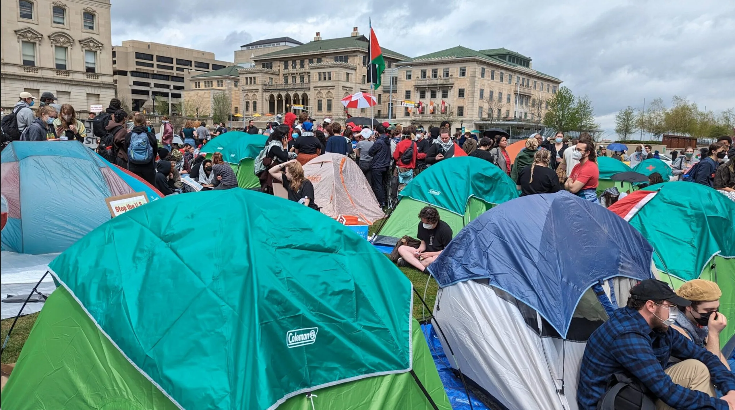 A large group of people gather among numerous tents on a grassy area in front of university buildings, with some holding flags and signs; the sky is cloudy.
