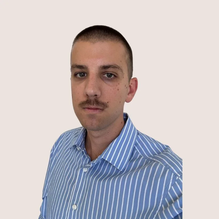 A man with short brown hair and a mustache, wearing a blue and white striped shirt, is looking at the camera against a plain light background.