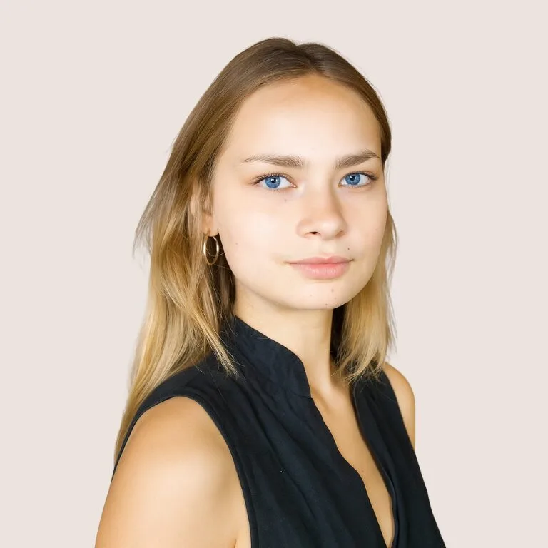 A young woman with straight blonde hair and blue eyes, wearing a sleeveless black top, looks at the camera with a neutral expression against a plain light background.