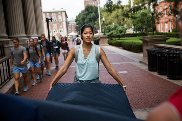 A young woman carrying a mattress outdoors on a college campus walkway, with other students and campus buildings visible in the background.