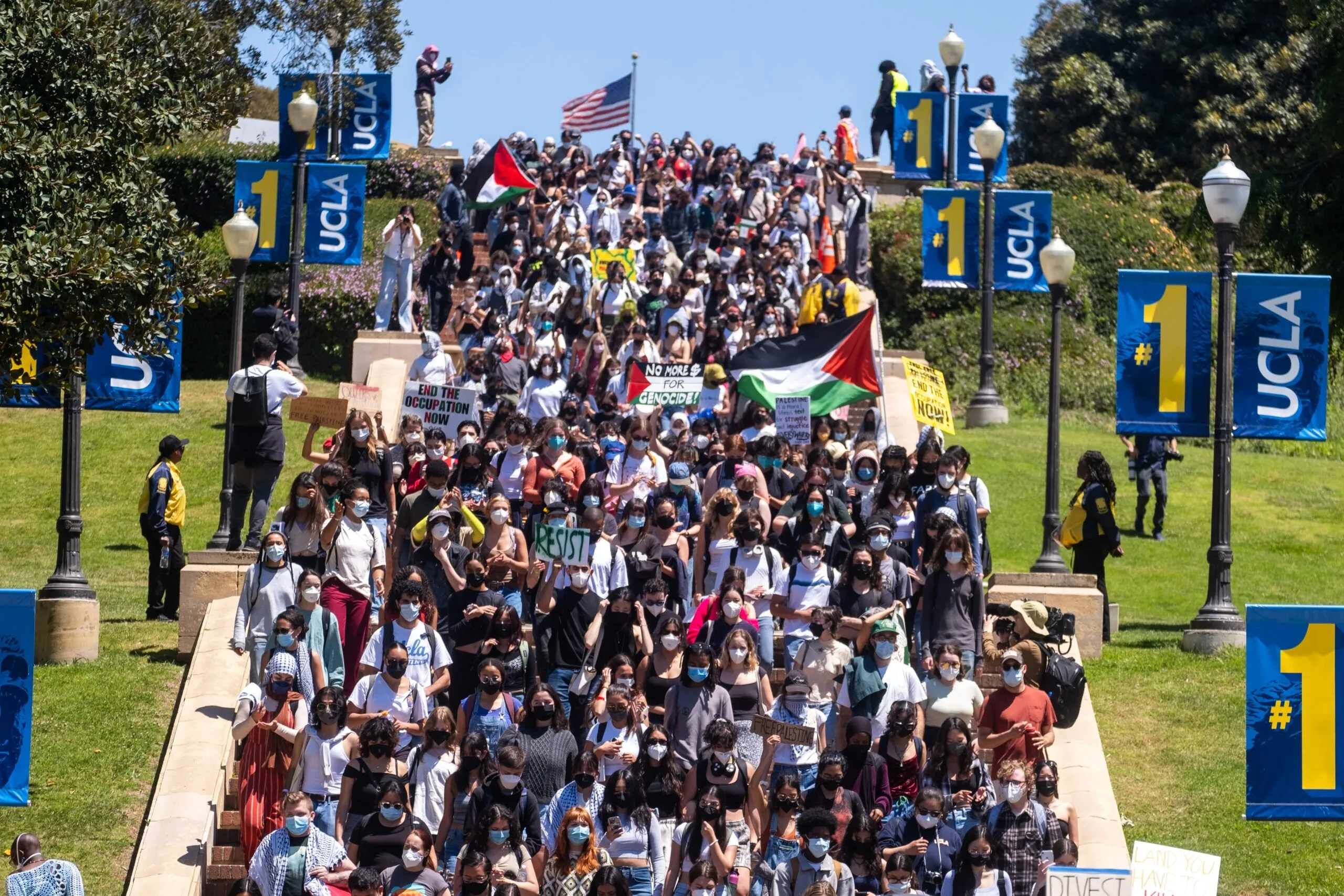 A large crowd of protesters holding signs and flags march down a staircase at UCLA, surrounded by blue banners with “UCLA” and “#1” on them; some people wear masks and an American flag waves in the background.