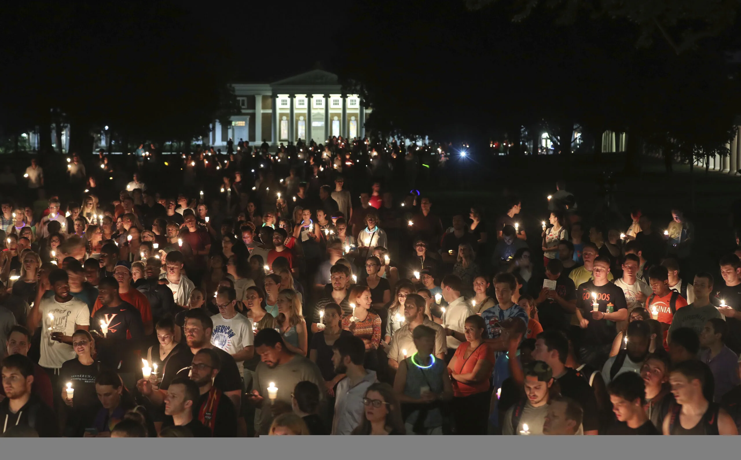 A large crowd gathers outdoors at night, holding lit candles in a vigil. The background features a well-lit building with columns, and the atmosphere appears solemn and reflective.