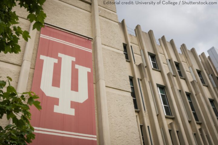 A large banner with the Indiana University logo hangs on the exterior of a beige building with tall windows and vertical architectural details. Green leaves are visible in the foreground.