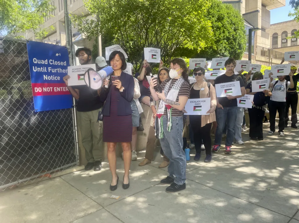 A group of people hold protest signs and listen to a speaker with a megaphone near a “Quad Closed Until Further Notice” sign on a sunny sidewalk, with trees and buildings in the background.