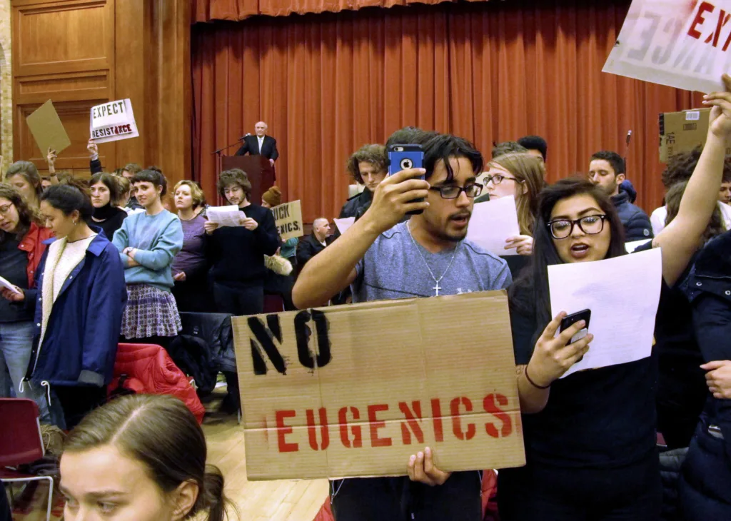 A diverse group of protesters holds up signs reading NO EUGENICS and others in a crowded room, while a person speaks at a podium in the background. Some protesters are taking photos or videos with their phones.