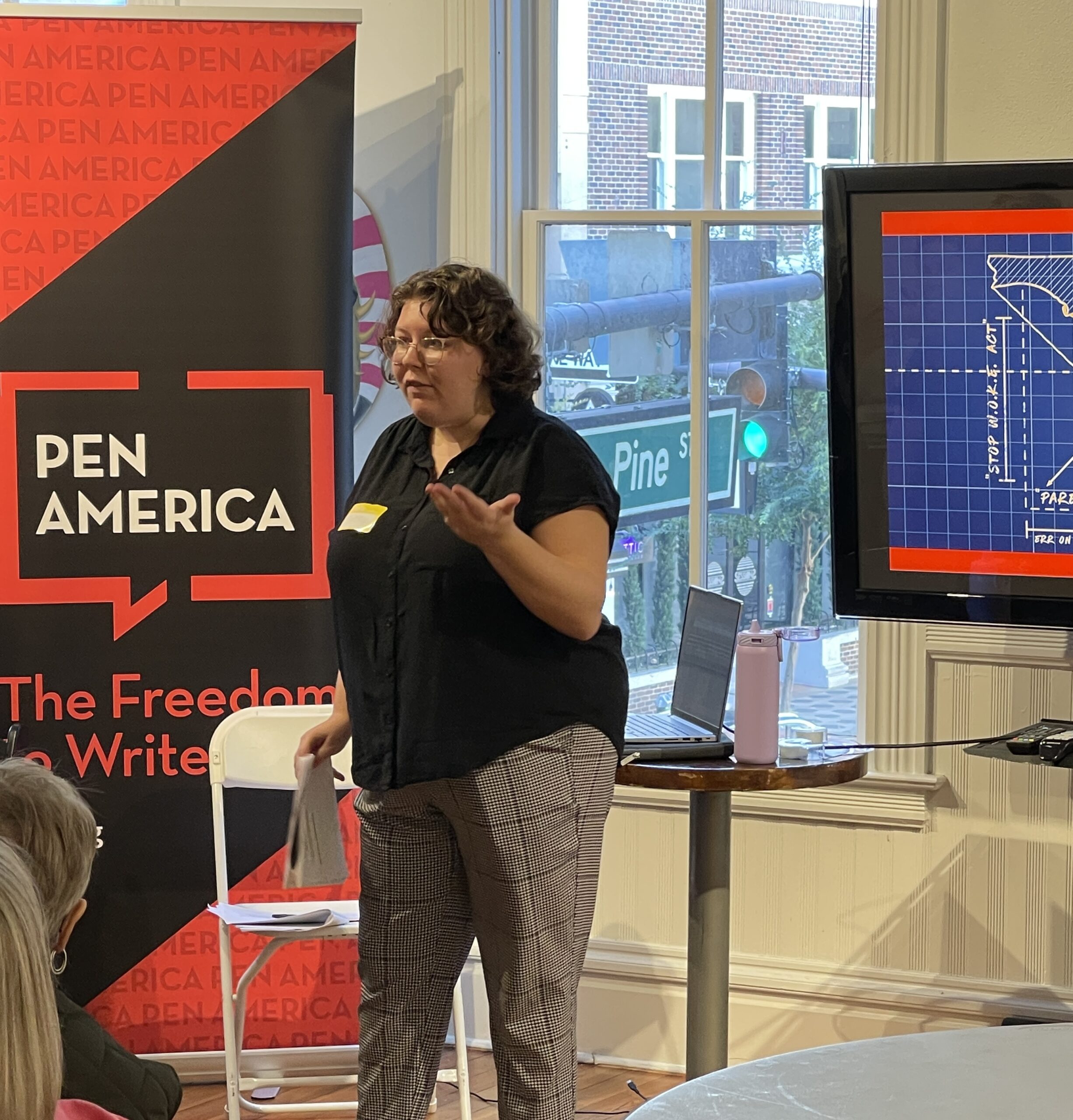 A woman stands in front of a PEN America banner and a screen with blueprints, speaking to an audience. She is gesturing with one hand and holding papers in the other. A window shows a street sign for Pine St outside.