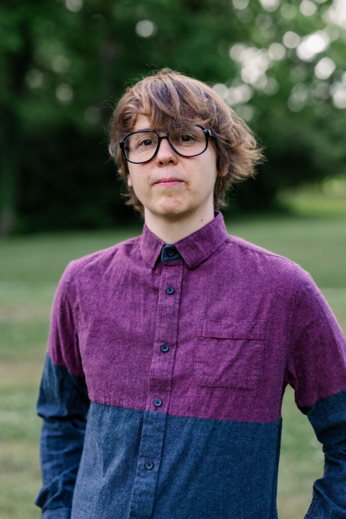 A young person with shaggy brown hair and black glasses stands outdoors, wearing a color-blocked button-up shirt in maroon and blue, with a neutral expression. Green trees and grass are blurred in the background.