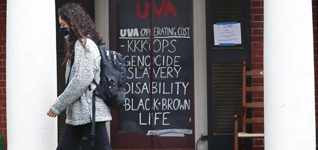A person wearing a backpack and mask walks past a dark door with a sign reading, “UVA operating cost: KKKops, genocide, slavery, disability, Black + Brown life,” and UVA is written in red above.