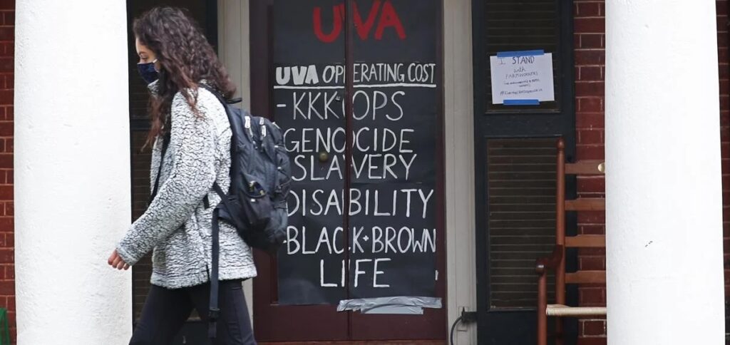 A person wearing a backpack and mask walks past a dark door with a sign reading, “UVA operating cost: KKKops, genocide, slavery, disability, Black + Brown life,” and UVA is written in red above.