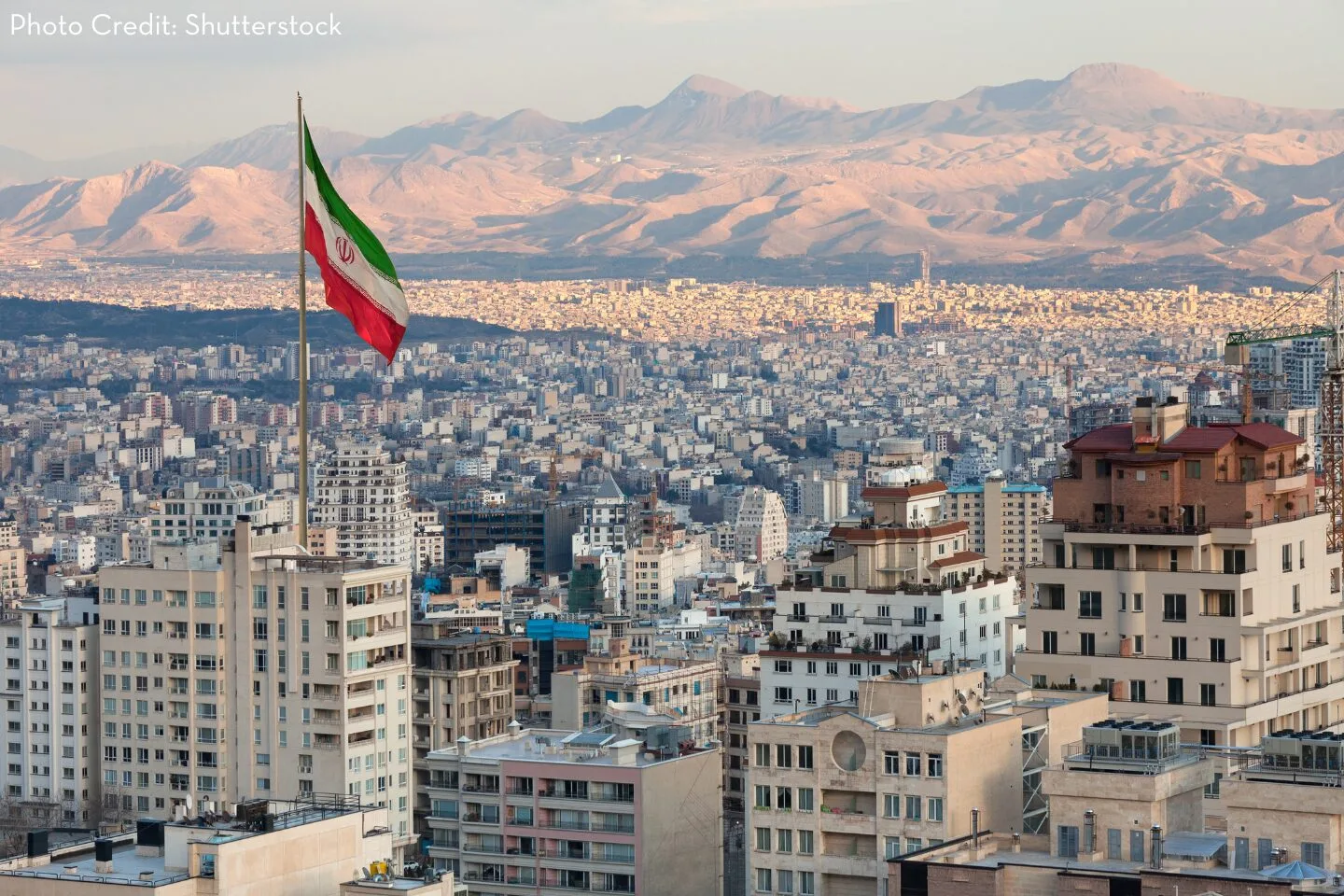 A large Iranian flag flies atop a building overlooking a densely packed cityscape of Tehran, with tall buildings in the foreground and mountains in the background under a hazy sky.