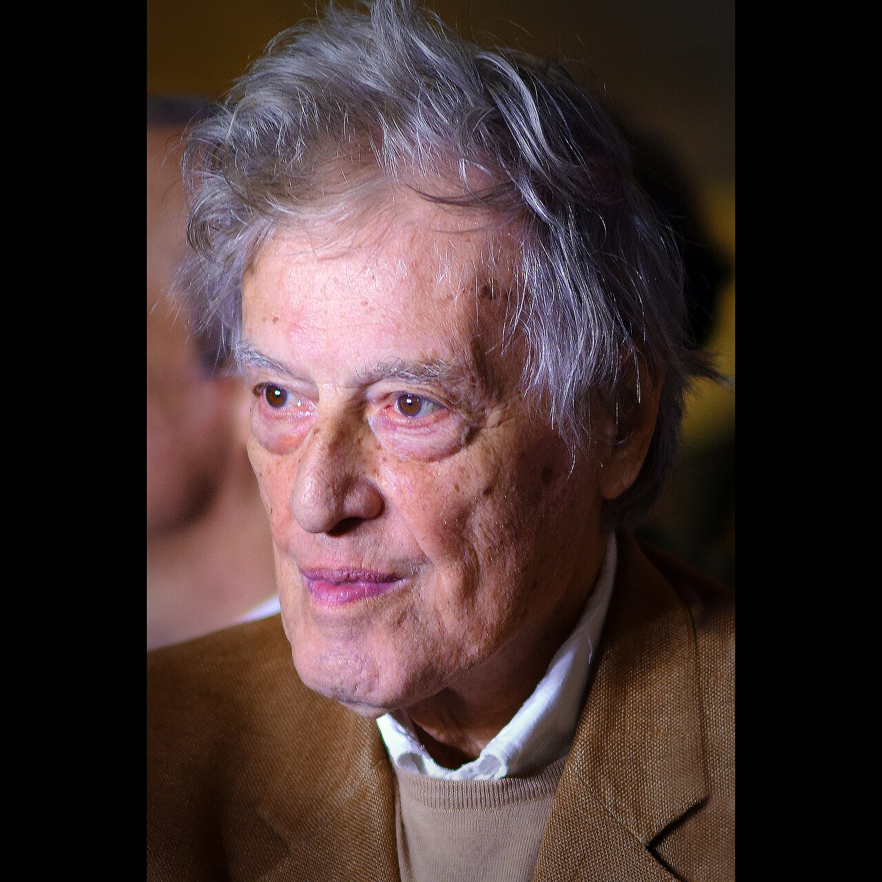 An elderly man with gray, tousled hair and a light brown jacket looks slightly to the side. The lighting highlights his face, showing a thoughtful expression and slight smile. The background is blurred.