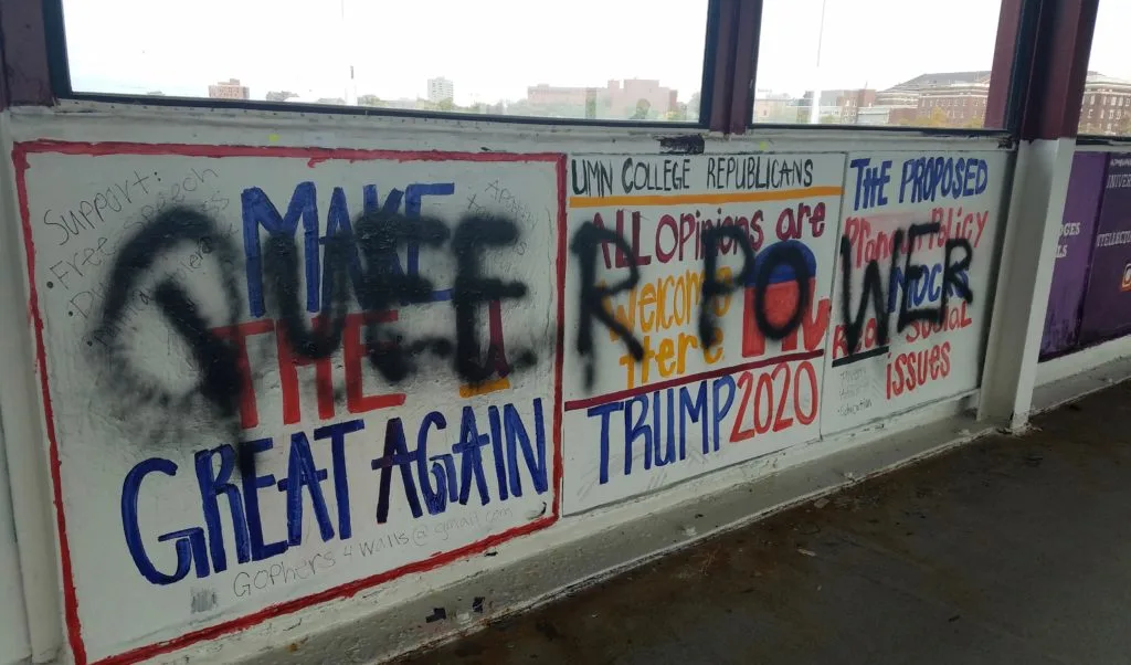A wall mural featuring Make America Great Again and pro-Trump slogans is defaced with black spray paint obscuring parts of the text. Messages about campus Republicans and political issues are partially visible.
