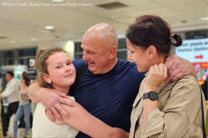 A smiling man embraces a young girl and a woman in an airport, sharing a joyful reunion. The background shows other people and airport signage.
