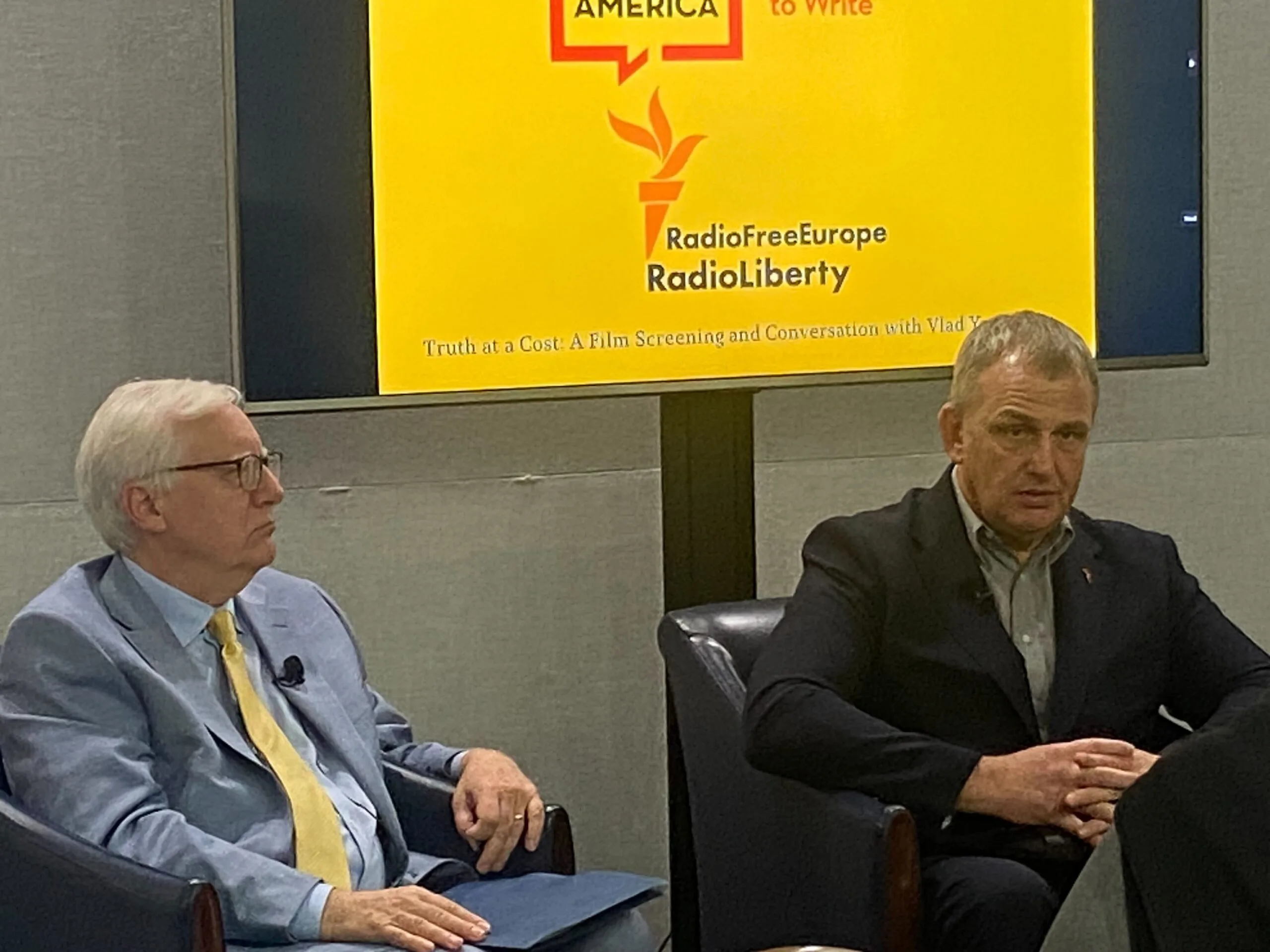 Two men in suits sit and speak in front of a large screen displaying the logos of PEN America and Radio Free Europe/Radio Liberty, during an event or discussion.