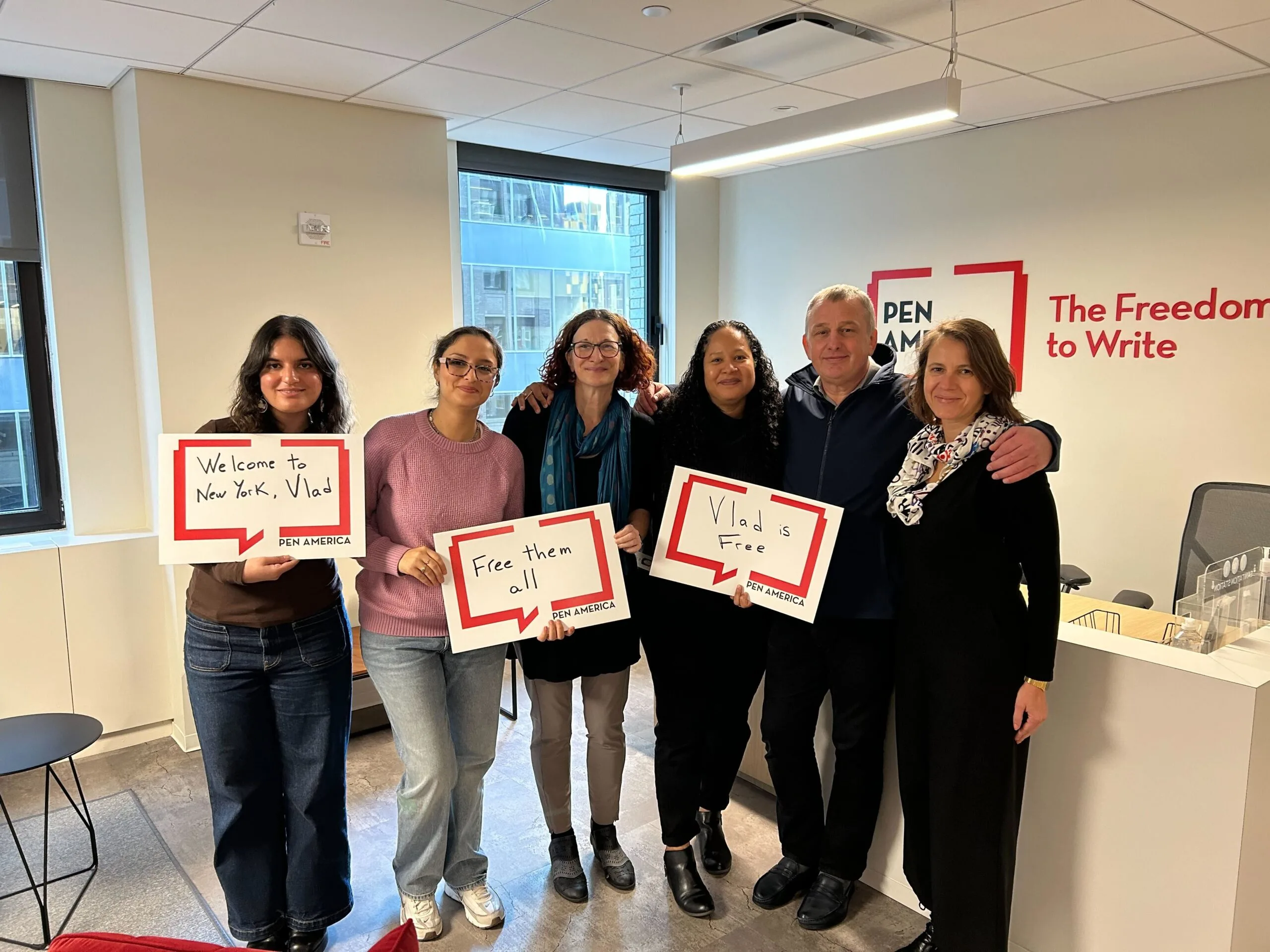 Six people stand together in an office, smiling and holding signs that read: Welcome to New York, Vlad, Free them all, and Vlads Free. A wall sign says PEN America: The Freedom to Write.