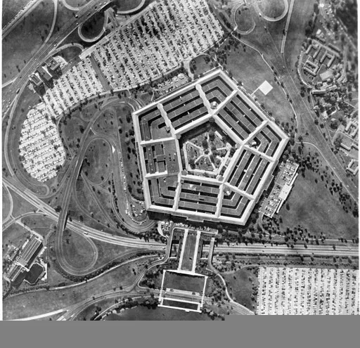 Aerial black-and-white photo of the Pentagon building, featuring its distinct five-sided shape, surrounded by roads, parking lots filled with cars, and green areas.