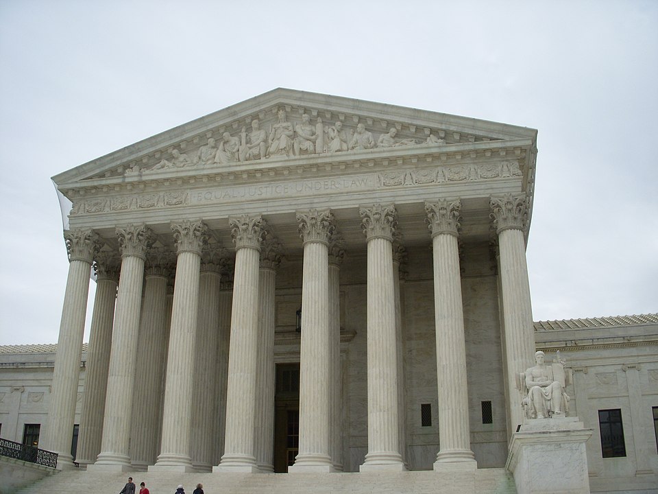 The image shows the front of the United States Supreme Court building in Washington, D.C., featuring large columns, detailed carvings on the pediment, and a statue on the right side of the steps under a cloudy sky.