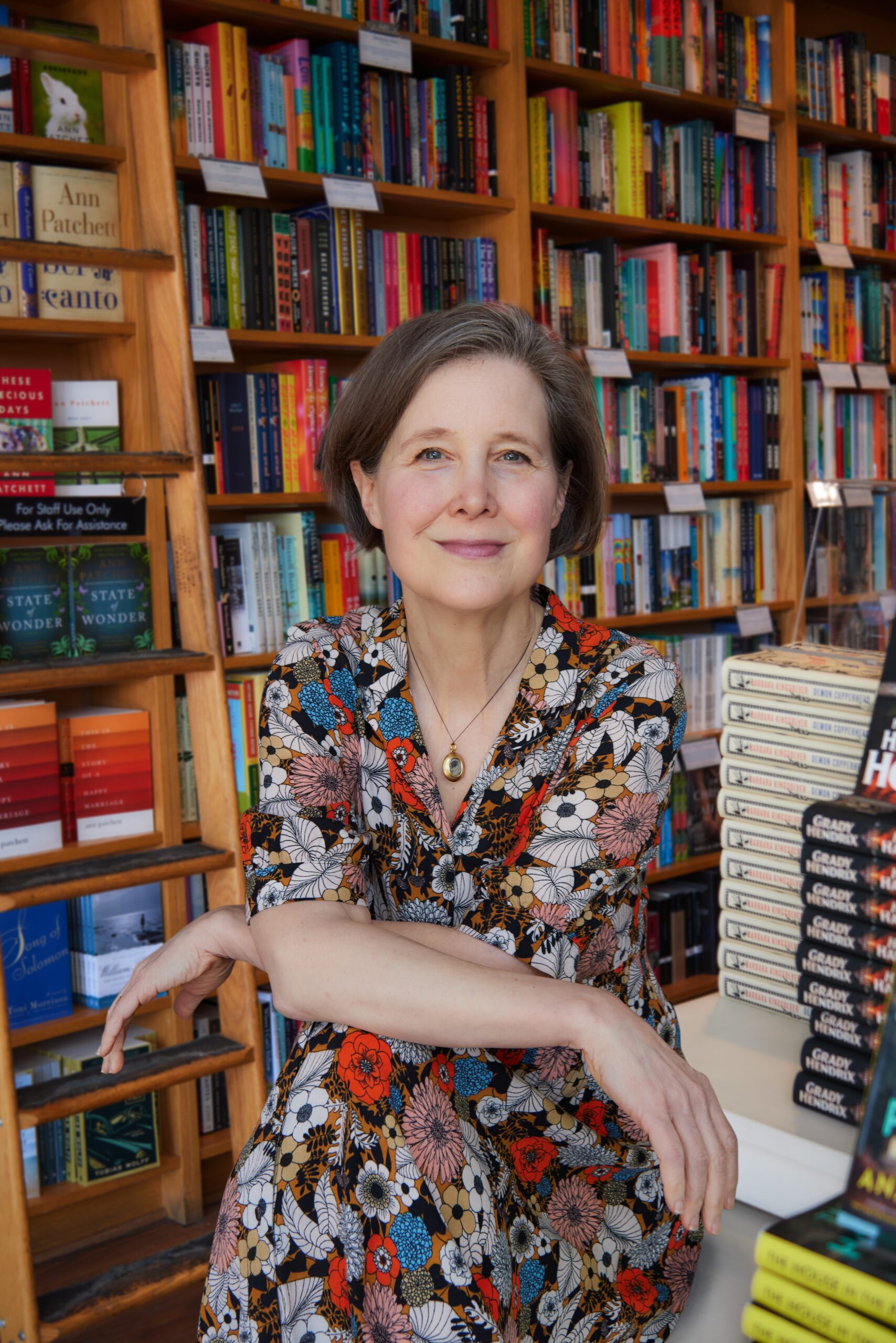 A woman with short brown hair wearing a floral dress sits with her arms crossed, smiling in front of shelves filled with colorful books inside a bookstore. A stack of books is visible beside her on the table.