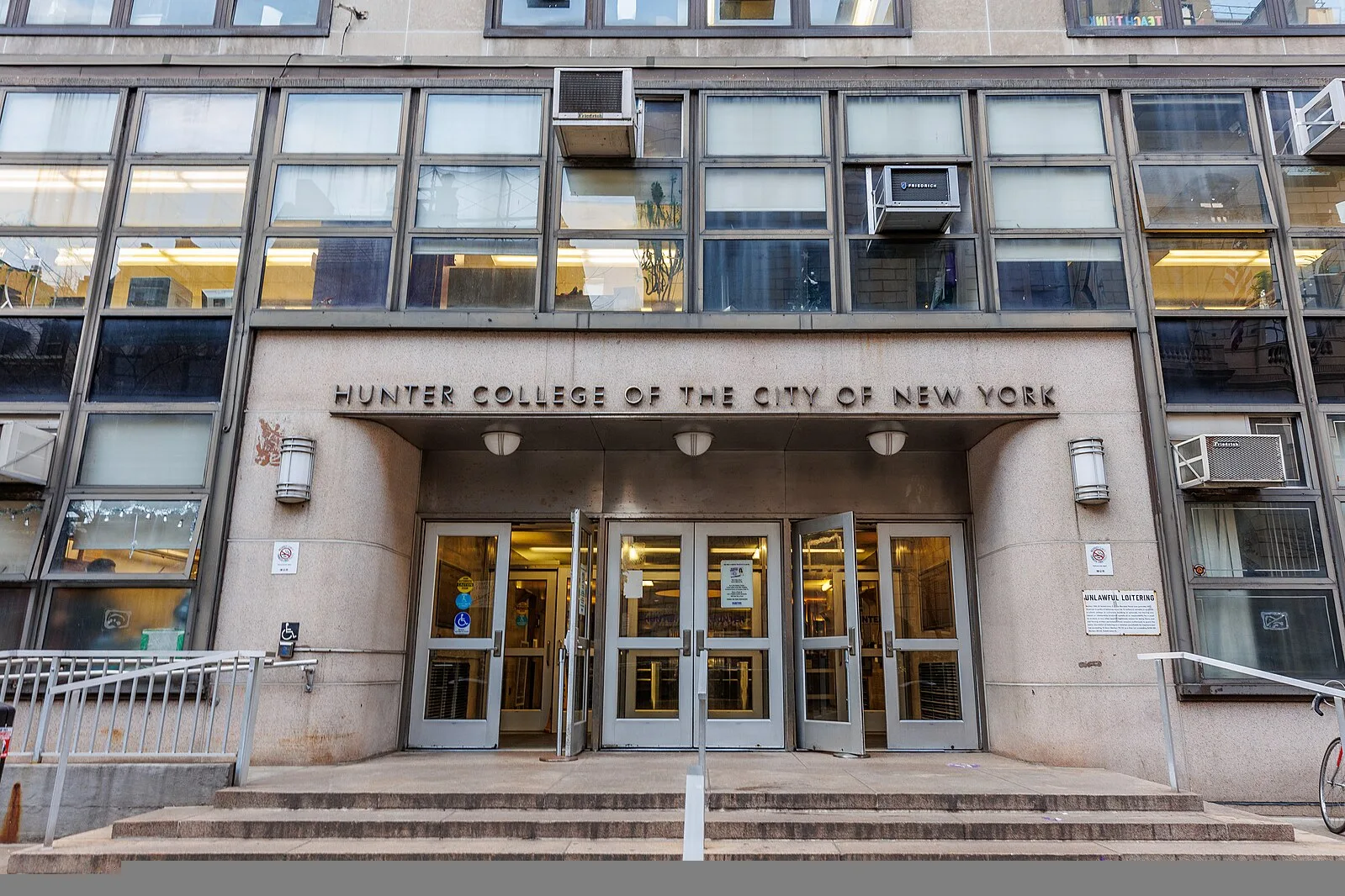 The entrance of Hunter College of the City of New York, featuring large glass windows, multiple doors, and signs above the doorway on a concrete building facade.