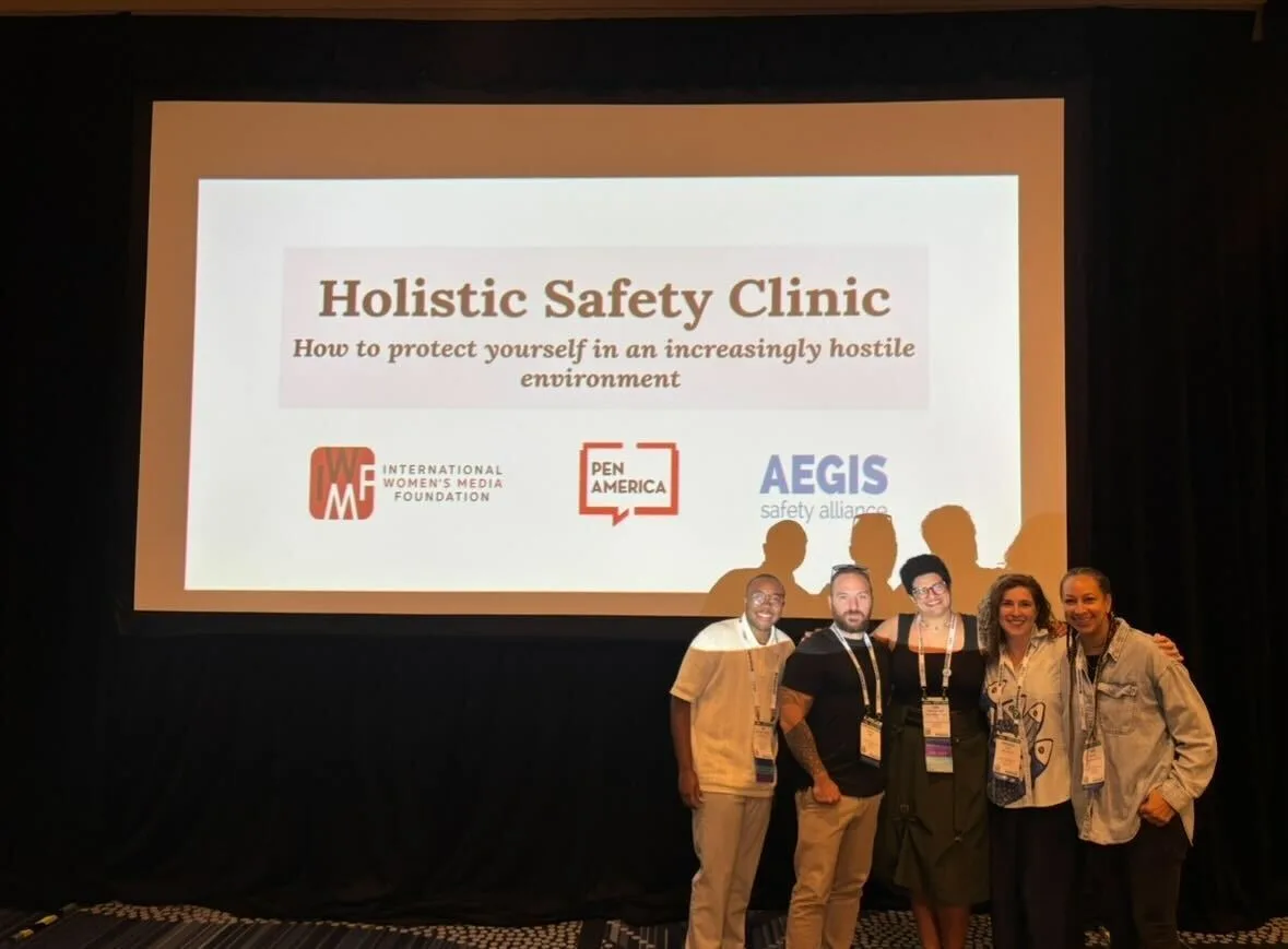 Five people stand in front of a screen displaying the title Holistic Safety Clinic: How to protect yourself in an increasingly hostile environment and logos for IWMF, PEN America, and AEGIS Safety Alliance.