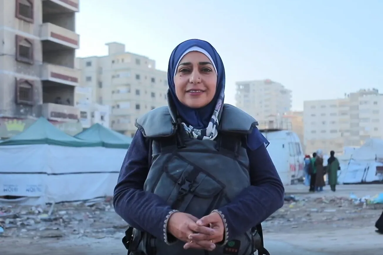 A woman wearing a hijab and protective vest stands smiling in front of tents and damaged buildings in an urban area, with people walking in the background.