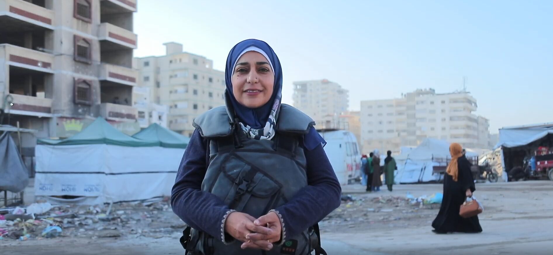 A woman wearing a hijab and protective vest stands smiling in front of tents and damaged buildings in an urban area, with people walking in the background.