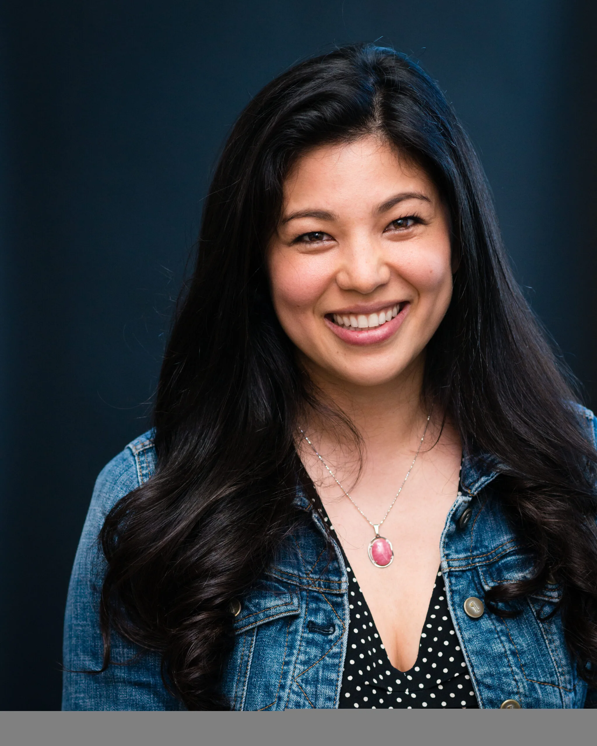 A woman with long dark hair smiles at the camera. She is wearing a denim jacket over a black blouse with white polka dots and a necklace with a pink pendant, standing against a dark, blurred background.