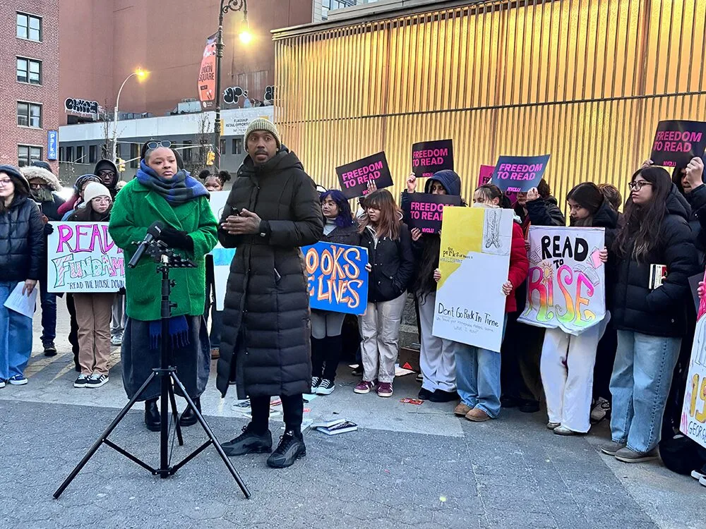 A group of people stand outside holding colorful signs that say “Freedom to Read” and “Read Rise.” Two speakers stand at a microphone in front of the group, with a building lit by yellow lights in the background.