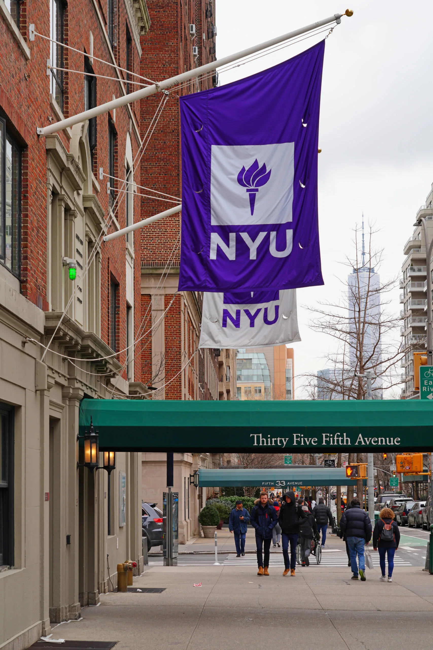 People walk on a sidewalk beneath large purple and white NYU flags outside a brick building with a green awning that reads Thirty Five Fifth Avenue in an urban city setting.