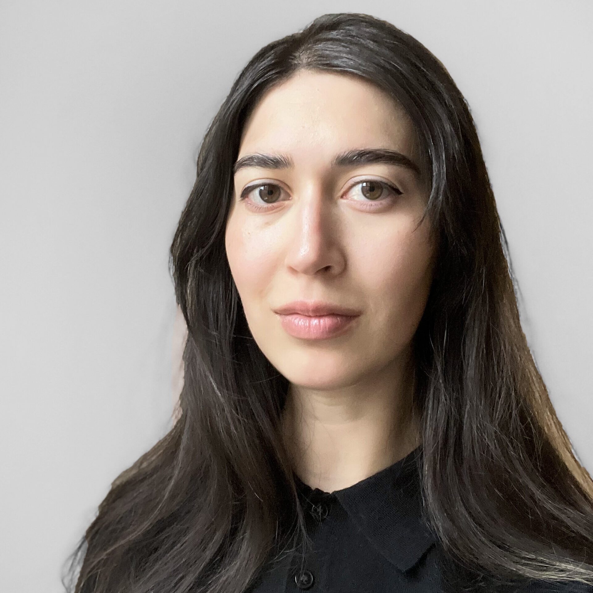 A woman with long dark hair, wearing a black collared shirt, stands against a plain light gray background, looking directly at the camera with a neutral expression.