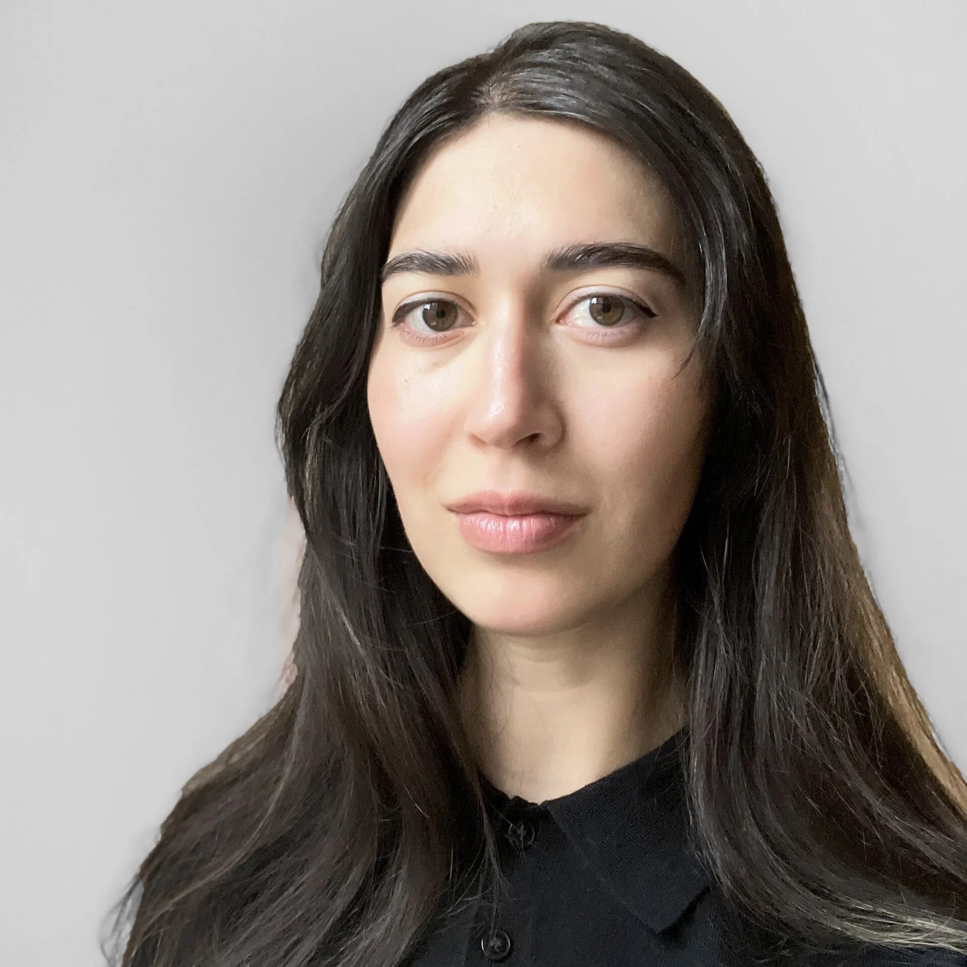 A woman with long dark hair, wearing a black collared shirt, stands against a plain light gray background, looking directly at the camera with a neutral expression.