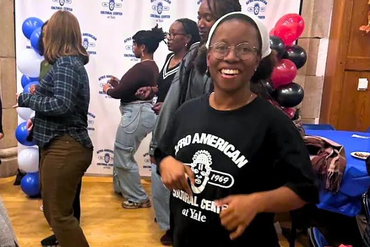 A smiling young woman wearing glasses and a Afro American Cultural Center at Yale shirt stands in the foreground at an indoor event, with other people and red, black, and white balloons in the background.